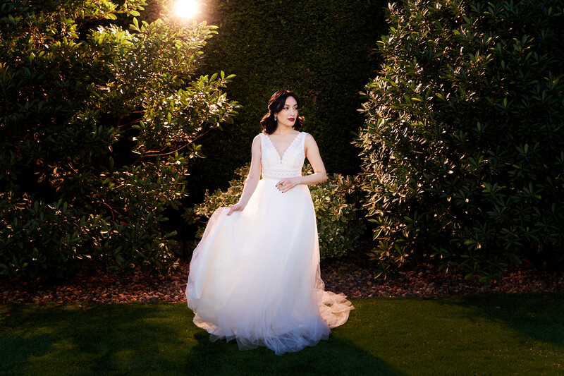 Woman in flowing white wedding dress standing on grass beneath illuminated green foliage at dusk.