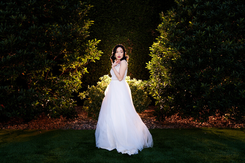 Woman in white flowing wedding gown standing on grass beneath illuminated hedge archway at night.