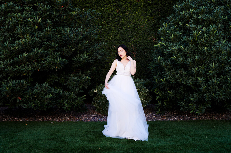 Woman in flowing white gown standing on grass against dense green hedge backdrop.