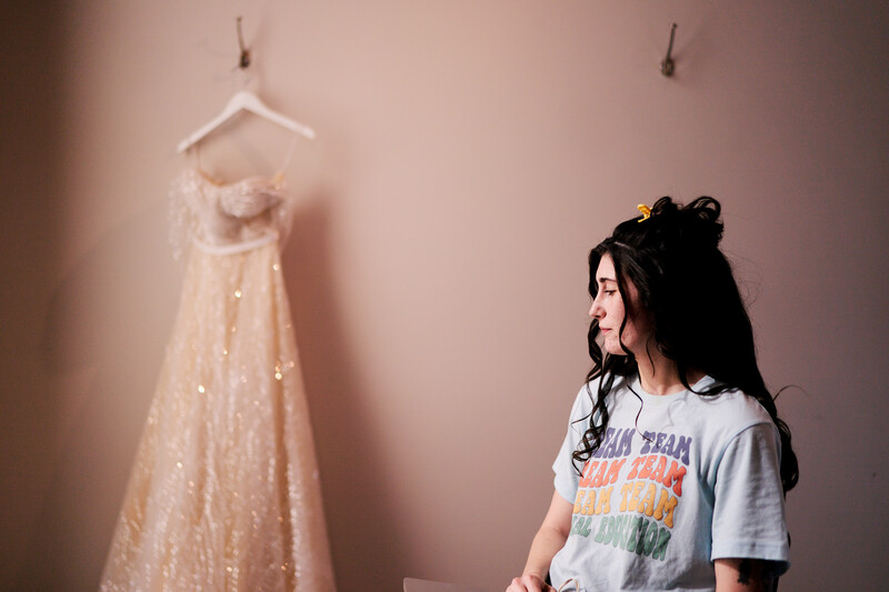 Woman in casual graphic t-shirt gazes toward an elegant beige gown displayed on a hanger against a pink wall.