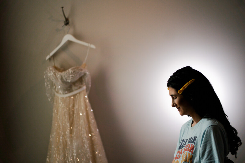 Woman in profile gazes at a shimmering beaded dress displayed on a hanger against a soft, backlit wall.