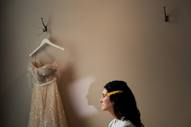 Woman in profile viewing a beaded gold dress on a white hanger against a neutral wall.
