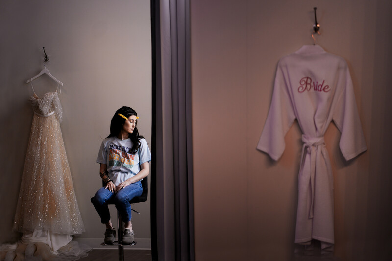 Woman seated between a beaded wedding dress and a pink silk robe with 'Bride' embroidered on the back.