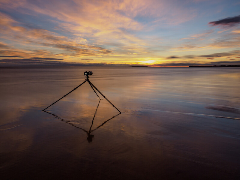 Tripod silhouetted against a golden sunset reflected in still shallow water.