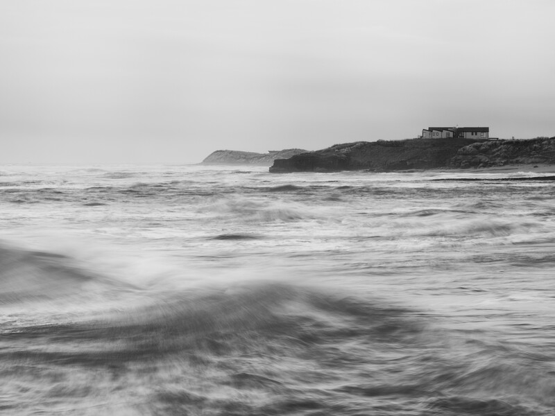 Stormy ocean waves crash on a beach with rocky headland and house visible on distant cliff.