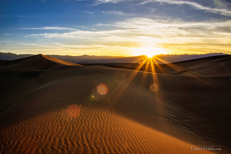 Golden sunlight streaks across sand dunes at sunrise, with mountains silhouetted against a bright sky.