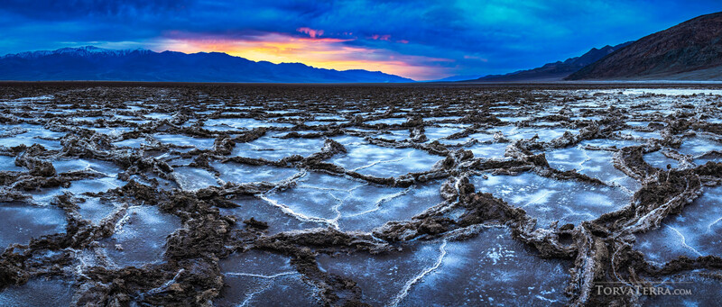 Cracked salt flats with shallow water pools under a dramatic twilight sky with mountains in the distance.