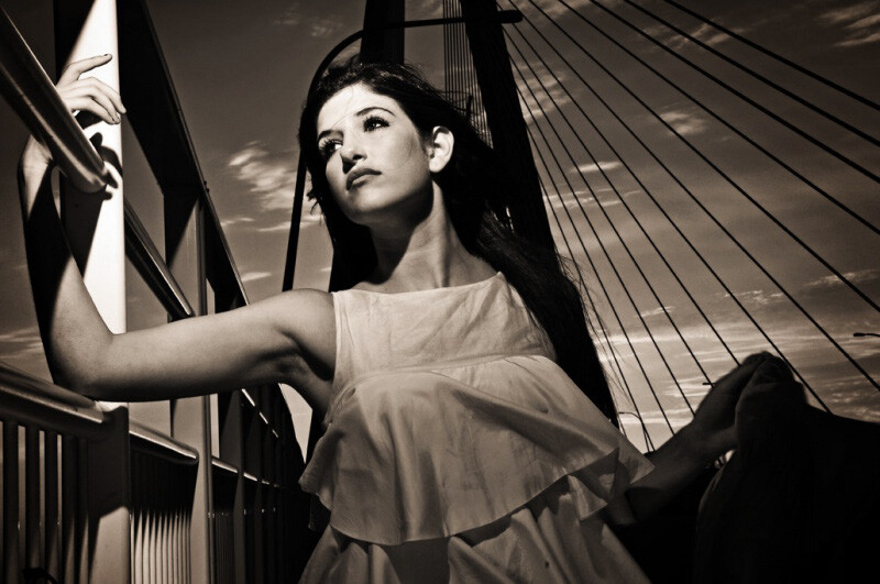 Woman in light dress posed against geometric bridge structure in high-contrast black and white photography.