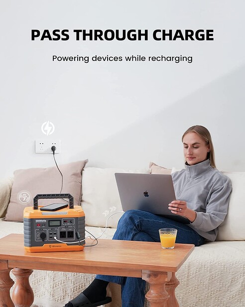 Woman working on laptop at wooden table with portable power station nearby, demonstrating pass-through charging capability.