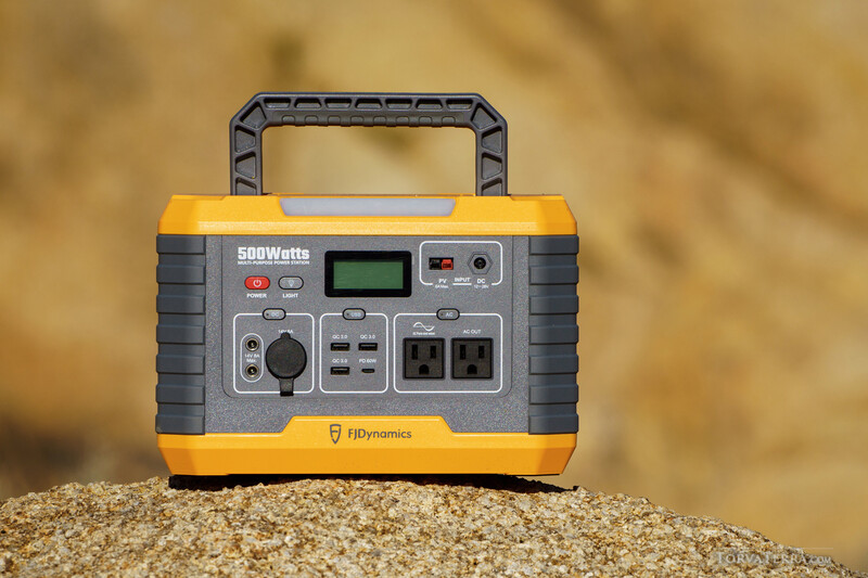 Portable power station with 500-watt capacity displayed on sandy surface against blurred background.