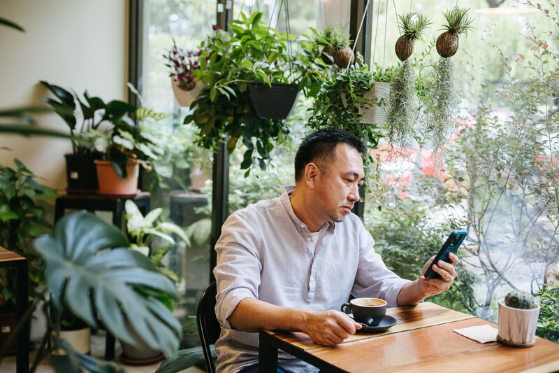 Man in light purple shirt working at wooden desk surrounded by hanging plants and potted greenery, holding smartphone.