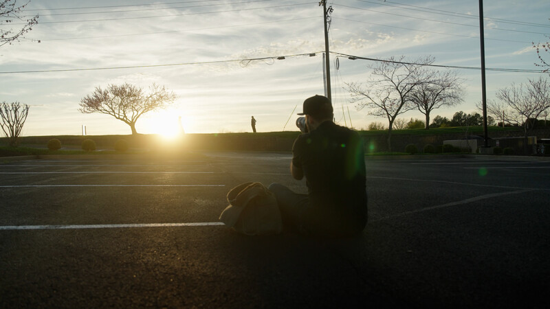 Silhouetted figure with camera photographing a sunset landscape with bare trees and power lines.