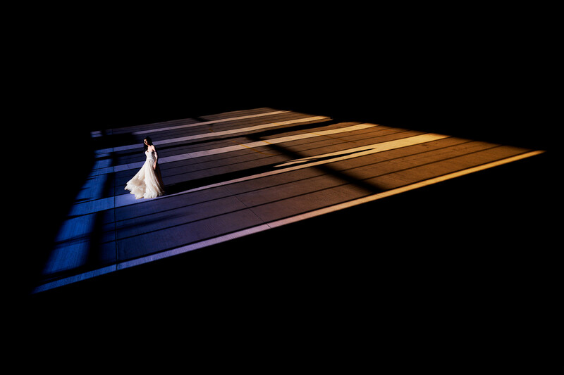 Bride in white wedding dress walking across dramatically lit wooden floor with blue and golden stage lighting against black background.