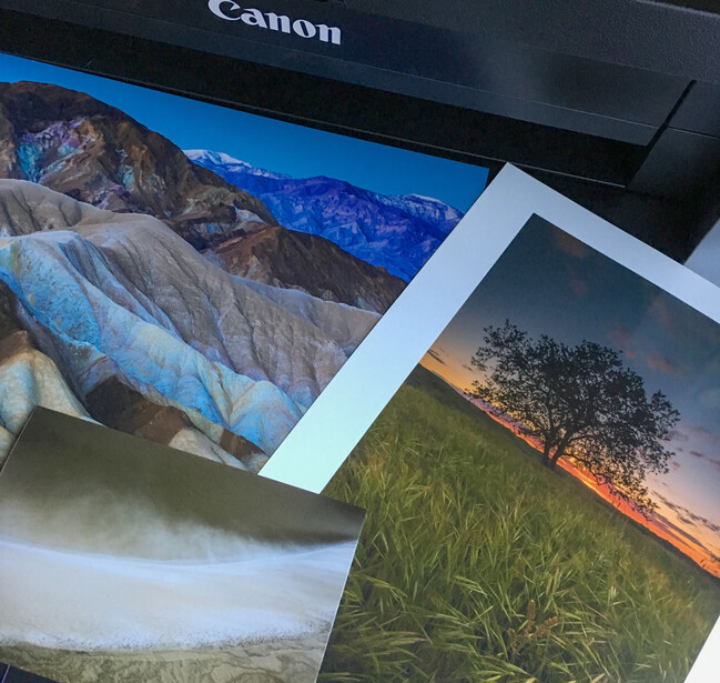 Printed landscape photographs fanned out on a Canon printer, showing colorful nature scenes with vibrant skies and terrain.