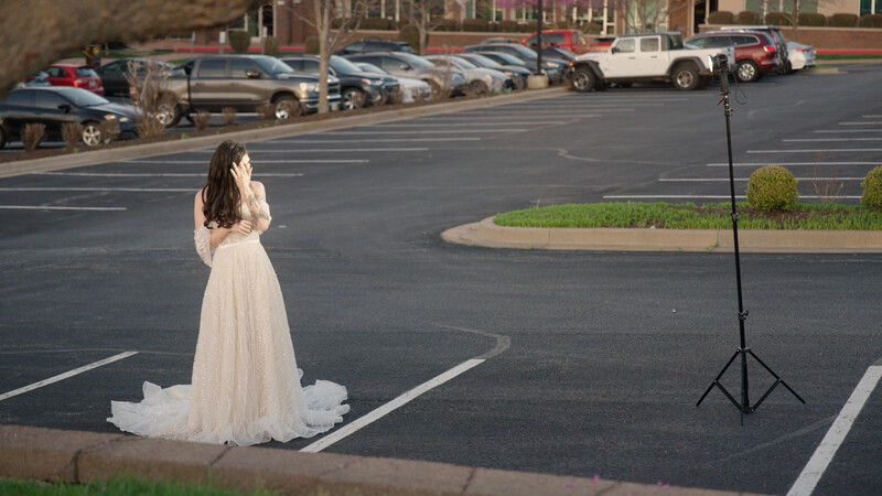 Bride in flowing white gown standing alone in empty parking lot with studio light on tripod.