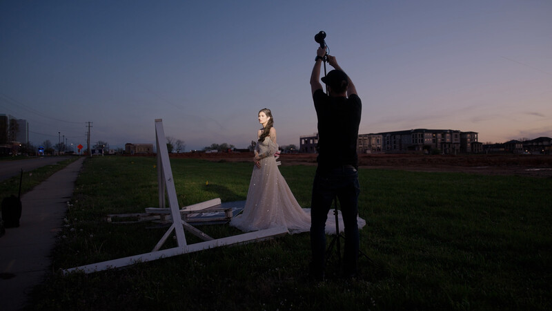 Bride in white wedding dress and groom in black suit holding up their joined hands at twilight in an open field.