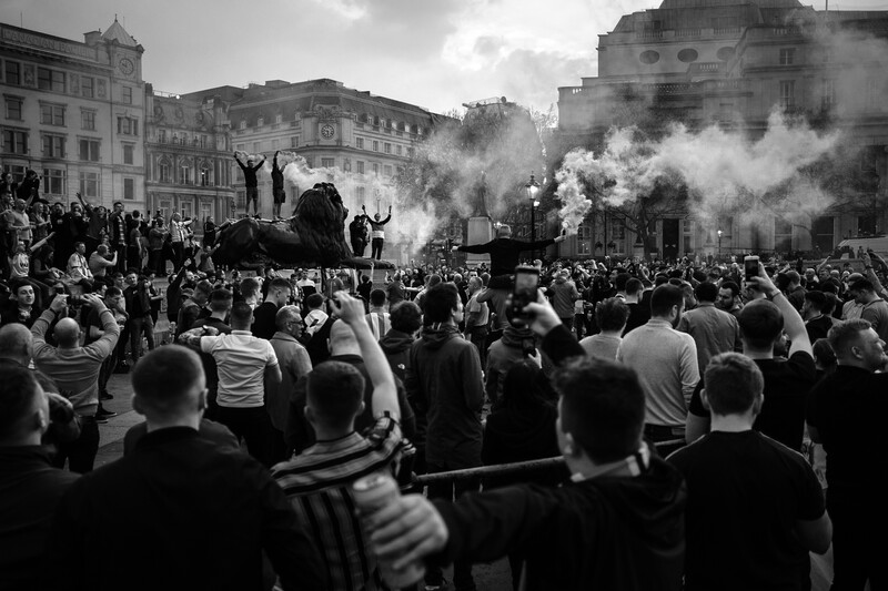 Dense crowd gathered in a city square with thick smoke billowing from multiple sources.