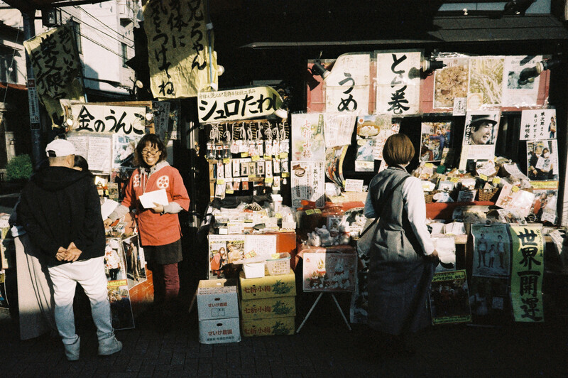 Nighttime street market stall displaying artwork, prints, and merchandise with vendors and customers browsing.