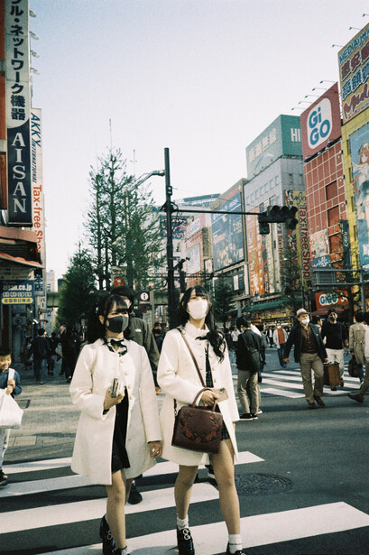 Two women in light-colored coats walking across a crosswalk in an urban commercial district with colorful signage.