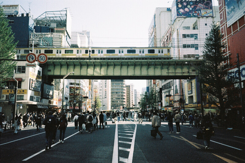 Elevated train crossing above a crowded urban street with tall buildings and pedestrians below.
