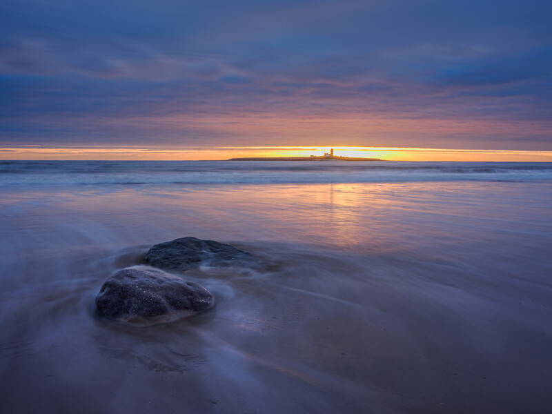 Rocky shoreline at sunset with smooth water reflections and golden sky illuminating distant structures.