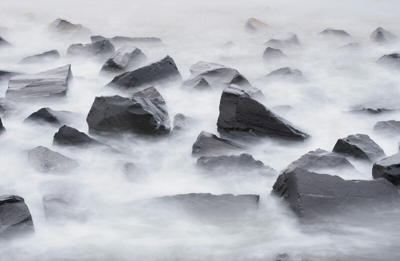 Rocky coastal boulders partially submerged in white foaming seawater with long exposure blur effect.