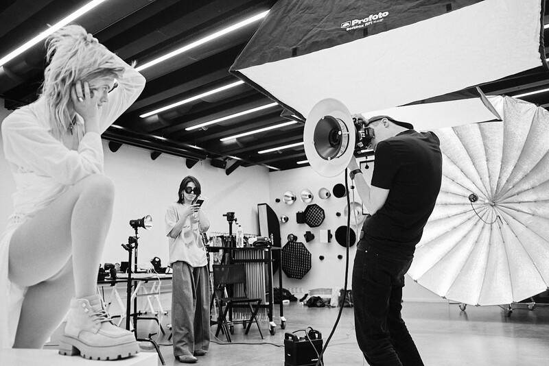 Professional studio photography setup with model, ring light, reflective umbrellas, and overhead lighting rig.