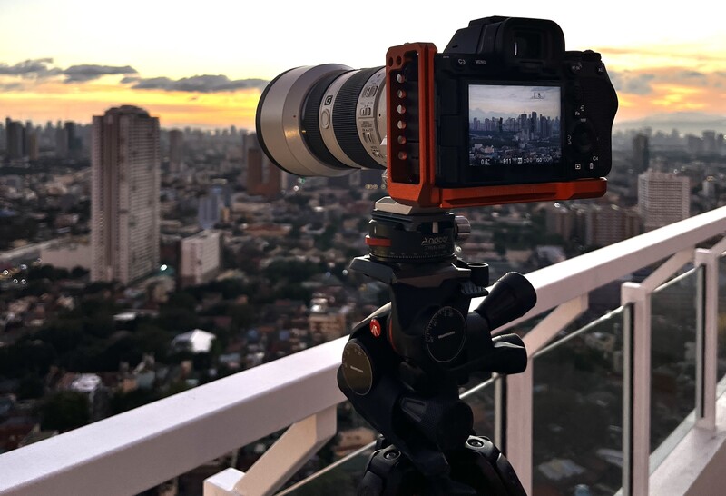 Camera with telephoto lens mounted on tripod overlooking city skyline at sunset.