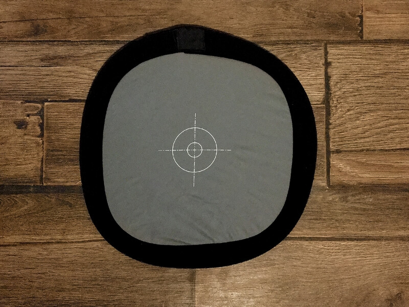 Gray photography reference card with white crosshair target overlay on wooden floor backdrop.