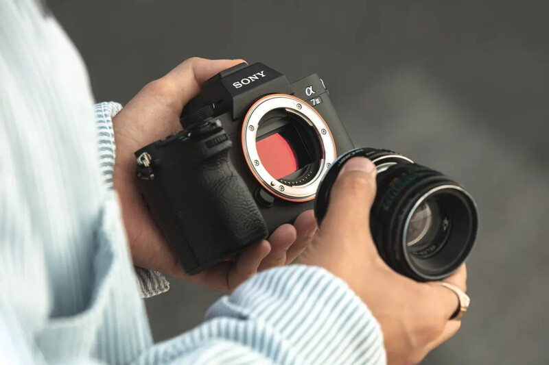 Photographer holding a Sony mirrorless camera body with mounted lens against neutral background.
