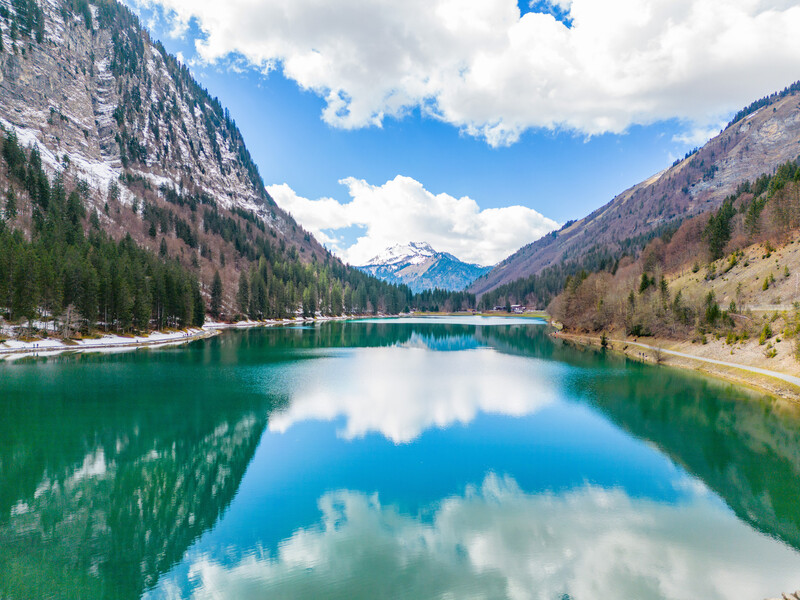 Alpine lake with mirror-like reflections of mountains, evergreen forest, and clouds under blue sky.
