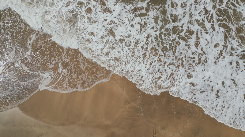 Aerial view of ocean waves breaking on sandy beach with white foam patterns.