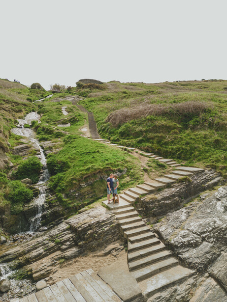 Hiker on wooden steps descending through green hillside landscape with stream.