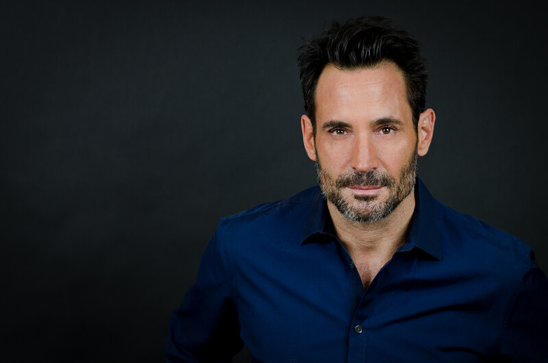 Man with dark hair and facial hair wearing a navy blue button-up shirt against a dark background.