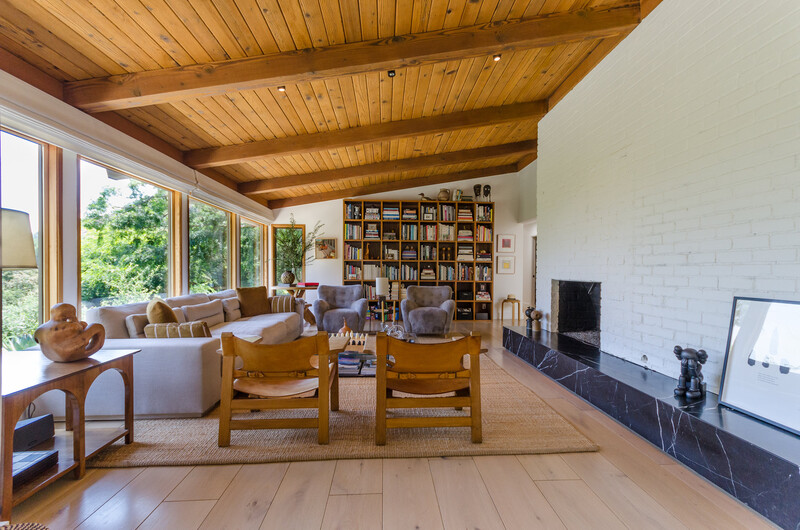 Modern living room with wooden exposed beam ceiling, large windows, mid-century wooden armchairs, and built-in bookshelves.