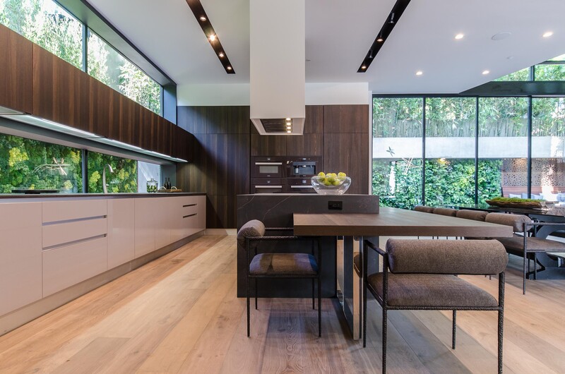 Modern kitchen and dining space with dark wood cabinetry, light wood flooring, and expansive windows overlooking trees.
