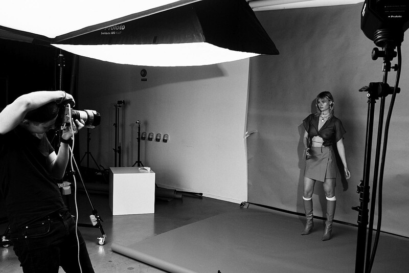 Photographer on left operating camera on tripod, facing model in athletic wear on studio platform under controlled lighting setup.