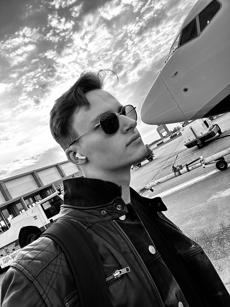 Person wearing sunglasses and black jacket photographed from below against sky and aircraft.