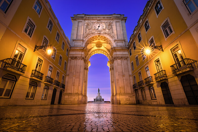 Illuminated archway framing a distant dome through symmetrical yellow buildings at dusk.
