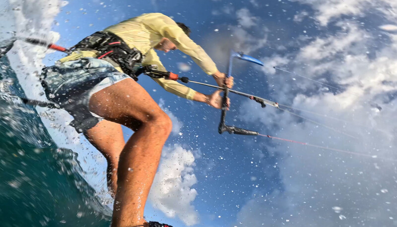Kitesurfer riding wave with yellow kite and board visible against blue sky with clouds.