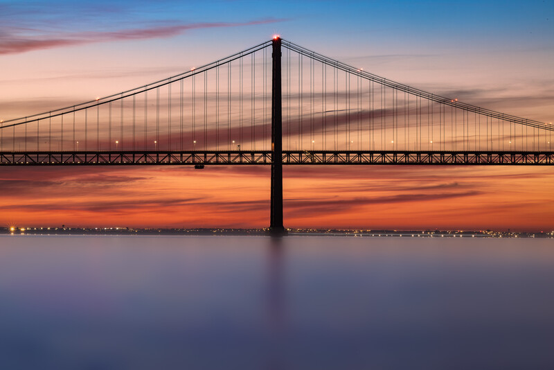 Suspension bridge silhouetted against a dramatic sunset sky with warm orange and pink hues reflecting on calm water below.