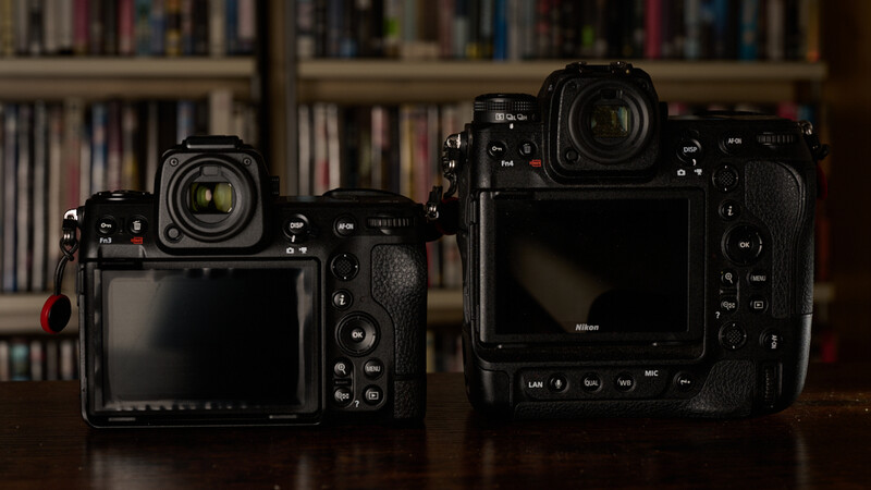 Two black mirrorless cameras side by side, the smaller model on the left and larger flagship model on the right, displayed against a blurred bookshelf background.