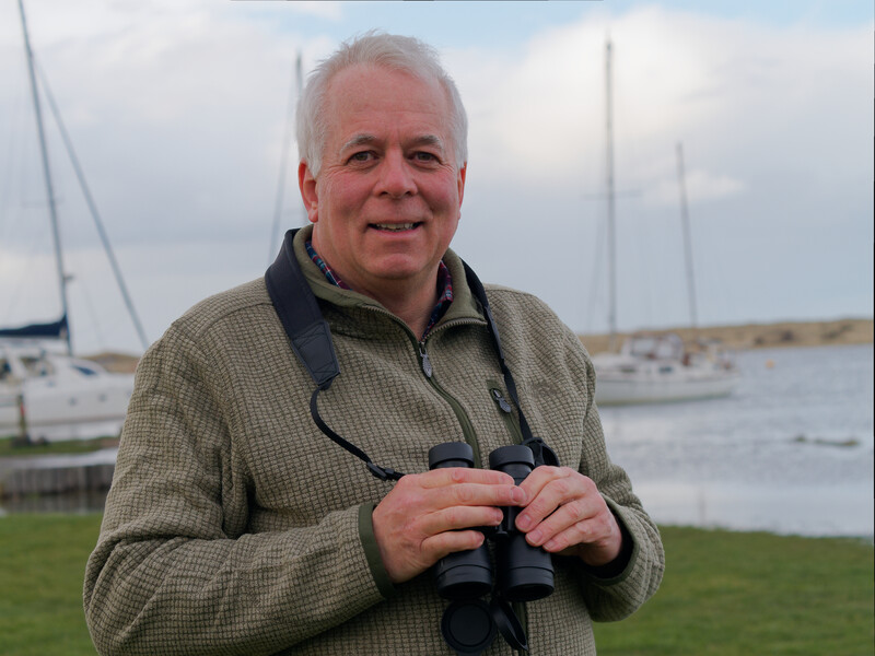 Man holding binoculars at a waterfront with sailboats moored in the background.