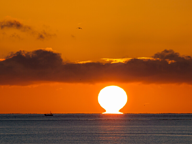 Sun touching the horizon at sunset over calm ocean water with dramatic orange sky and clouds.
