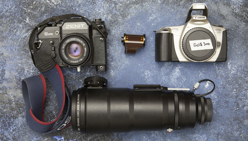 Collection of vintage film cameras and a telephoto lens arranged on a textured gray surface.