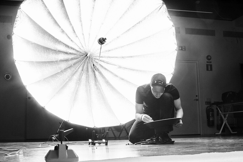 Photographer crouched beside large parachute reflector on studio floor during lighting setup.