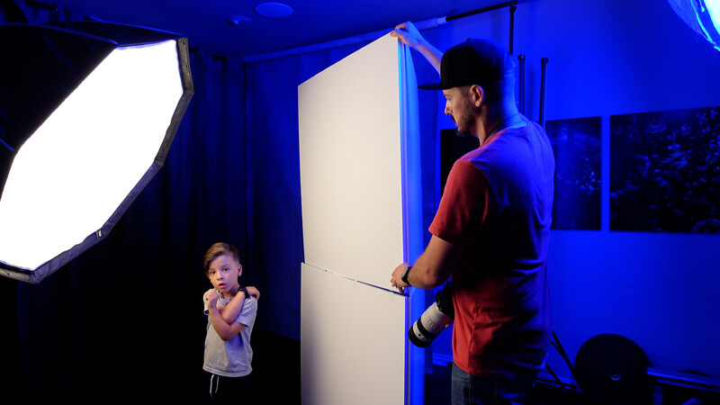 Photographer adjusting a large white reflector panel while positioning a young child subject in a studio lit with blue overhead lights.