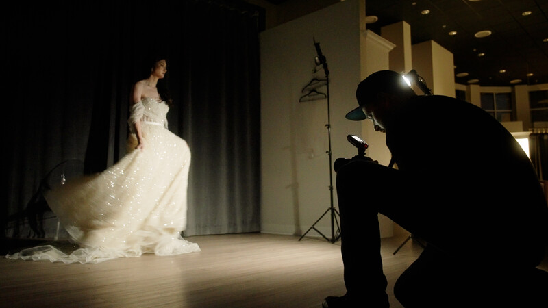 Photographer with camera on tripod shooting a bride in an elaborate white gown during a studio fashion shoot.