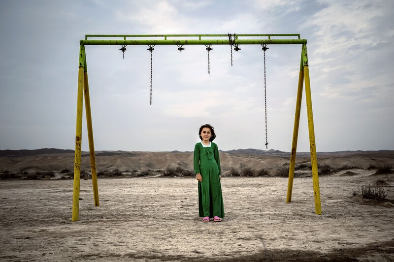 Woman in green dress standing beneath an empty swing set in a desert landscape.