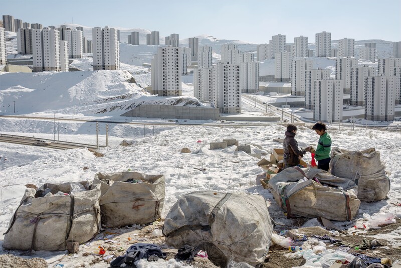 Two people sitting on rocks overlooking a sprawling residential cityscape with snow-covered ground and mountains in the distance.